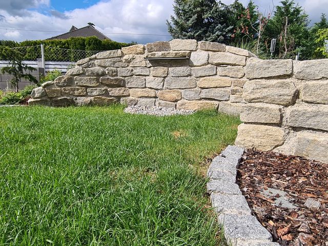 Eine Gartenlandschaft mit einer niedrigen, gebogenen Steinmauer, die als Terrassenbegrenzung dient. Der Garten besteht aus einer grünen Rasenfläche und einem angrenzenden Beet mit gelben und grünen Pflanzen. Im Vordergrund rechts ist ein Bereich mit Rindenmulch und einer kleinen Pflanze zu sehen. Im Hintergrund sind ein Maschendrahtzaun und benachbarte Häuser mit Gärten zu erkennen. Der Himmel ist teilweise bewölkt, und die Umgebung wirkt gepflegt und ruhig.