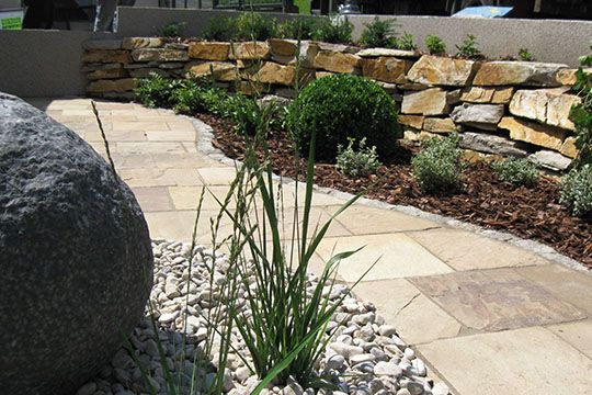 Ein Steingarten mit einem Weg, der von Pflanzen und Büschen gesäumt ist, führt zu einem Gebäude.