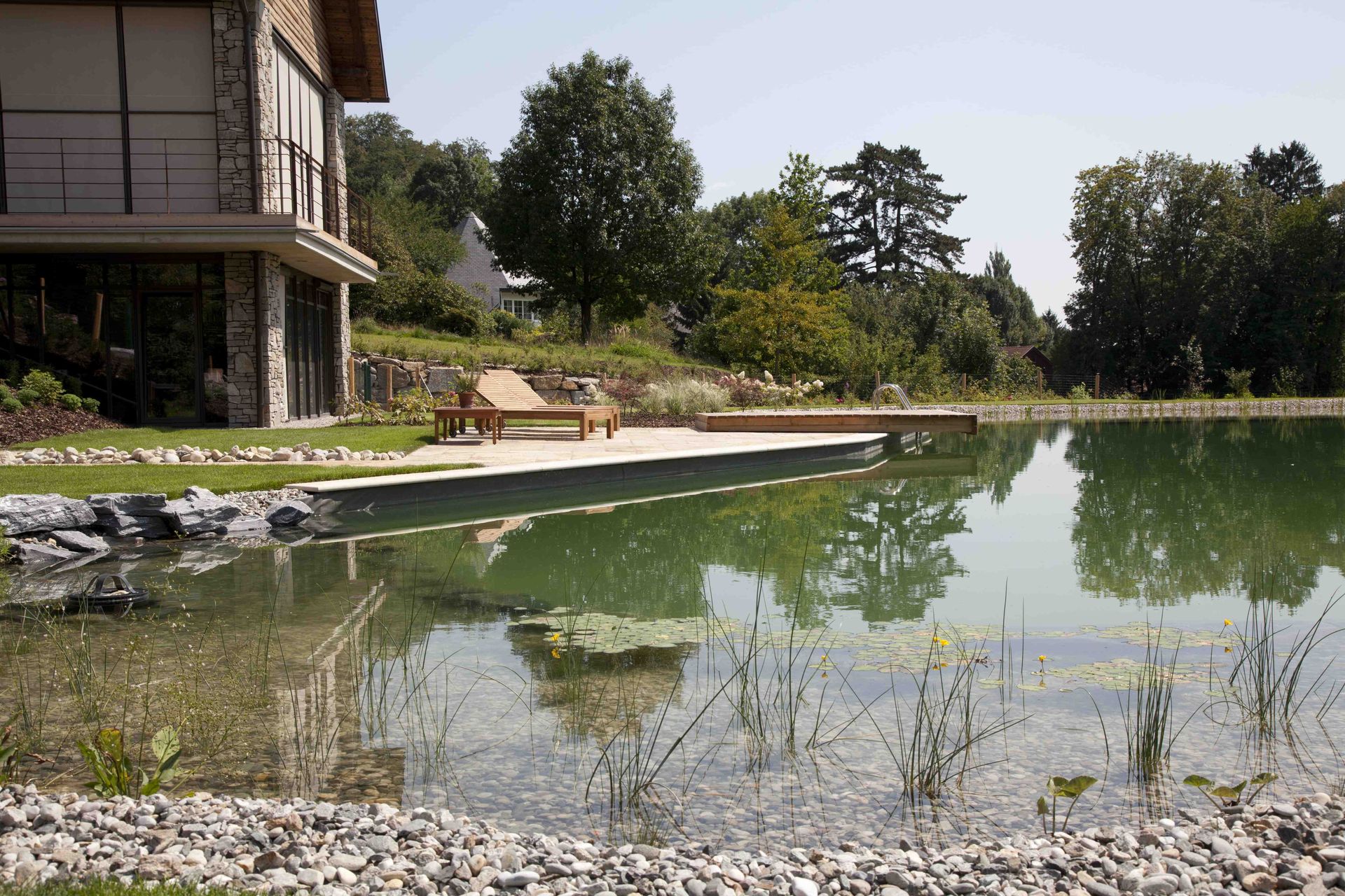 Schöner Naturpool, umgeben von Steinen und Gras, mit einer modernen Holzliege im Hintergrund.