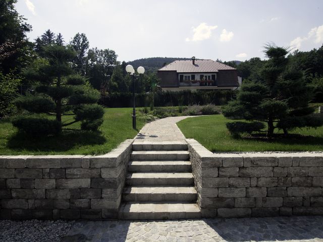 Steintreppe führt zu einem gepflasterten Weg, der durch einen gepflegten Garten mit geschnittenen Bäumen und einer Laterne verläuft; im Hintergrund ein Haus mit rotem Ziegeldach und Balkon, umgeben von grünen Bäumen und bewaldeten Hügeln.