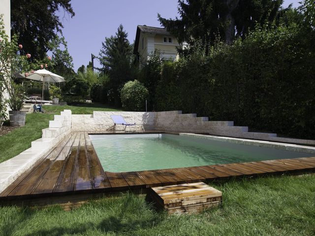 Ein rechteckiger Naturpool mit einer hölzernen Umrandung und treppenartigen Steinstufen an der Seite, umgeben von grüner Vegetation und einem gepflegten Garten. Im Hintergrund sind eine Gartenbank, ein Tisch mit Sonnenschirm und ein gelbes Haus sichtbar, was dem Bild eine gemütliche und einladende Atmosphäre verleiht.