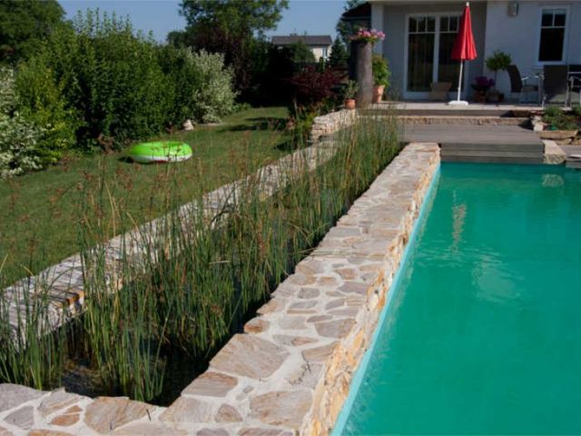Ein Pool mit Steinmauer und bepflanztem Rand, mit einem grünen Luftkissen und einem roten Sonnenschirm im Hintergrund.