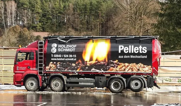 Pellets LKW in roter Farbe und Logo von Holzhof Schmidt. Roter Lkw der Firma Holzhof Schmidt mit großflächiger Werbung für Holzpellets. Auf der Plane sind brennende Pellets abgebildet sowie die Aufschrift ‚Pellets aus dem Wechselland‘. Der Lkw steht auf einem nassen Hof, im Hintergrund sind Holzstapel und Bäume zu sehen.
