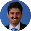 Smiling man in a suit and blue tie against a blue background.