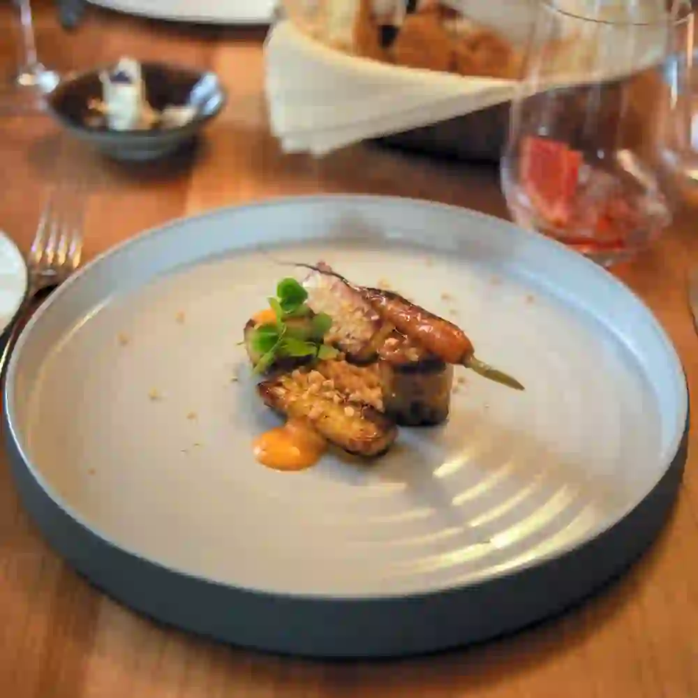 This picture shows an elegantly presented dish on a simple blue plate. The culinary composition with carefully placed ingredients and a harmonious sauce reflects modern, creative cuisine. The focus on the food in front of a blurred restaurant interior suggests an intimate and high-quality dining experience.