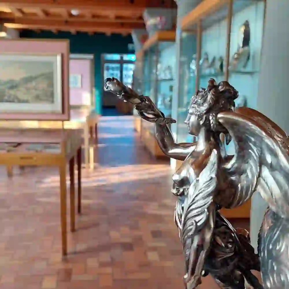 L'image montre l'intérieur du Musée du tir à Berne avec une vitrine en verre et plusieurs œuvres d'art exposées. Au premier plan se trouve une sculpture très détaillée d'une femme ailée tenant un objet dans la main. En arrière-plan, on peut voir d'autres objets exposés et œuvres d'art dans des vitrines ainsi qu'un tableau sur le mur. La salle a une charpente rustique en bois et est bien éclairée.