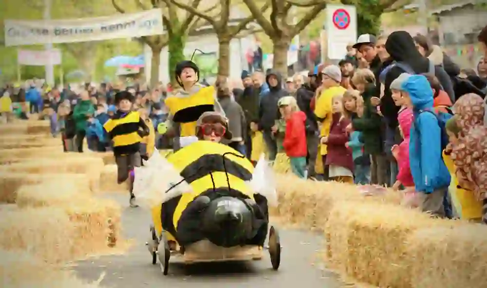 L'immagine mostra la gara di soapbox delle Grandi Giornate di Corsa Bernesi. Un veicolo ape dal design fantasioso corre lungo la pista, accompagnato da corridori in costume. La folla applaude, i bambini sono stupiti: entusiasmo puro, creatività e febbre da corsa sono nell'aria!