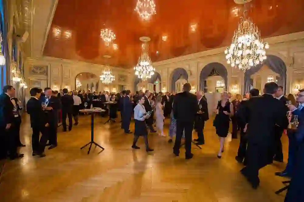Das Foto zeigt eine elegante Empfangshalle mit Menschen, die sich bei einer Veranstaltung unterhalten. Kristallleuchter hängen von der Decke, und die Stimmung ist gesellig und festlich.