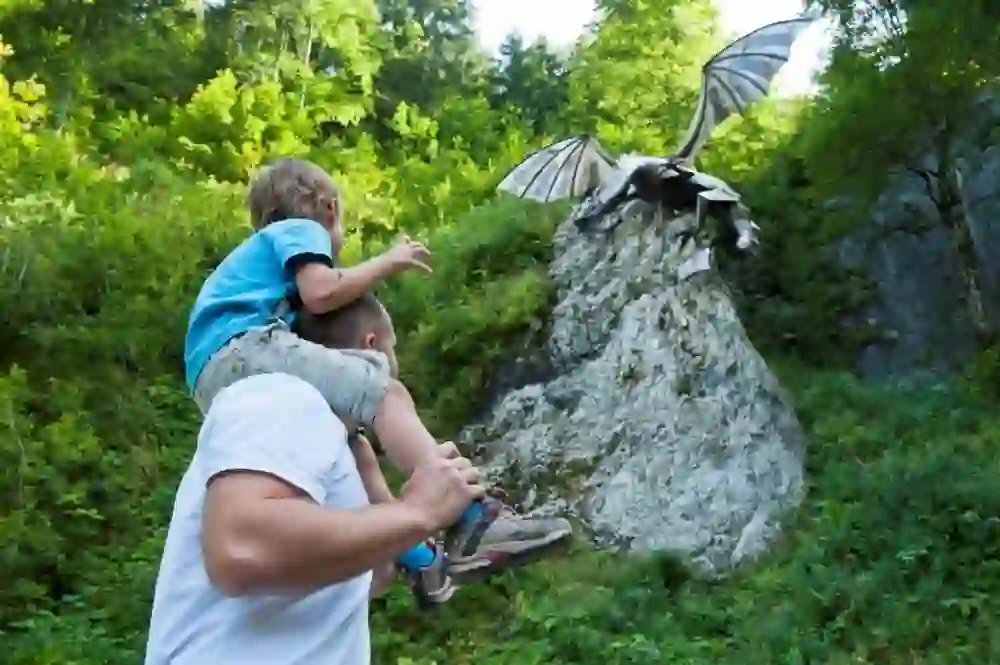 Auf dem Bild ist ein Mann zu sehen, der ein Kind auf seinen Schultern trägt. Sie scheinen eine fantastische Szene zu betrachten, in der ein Drache auf einem Felsen landet. Die Umgebung ist idyllisch und grün, und die Aufregung und das Staunen des Kindes sind spürbar. Es ist ein Moment des Wunderns und der Fantasie.
