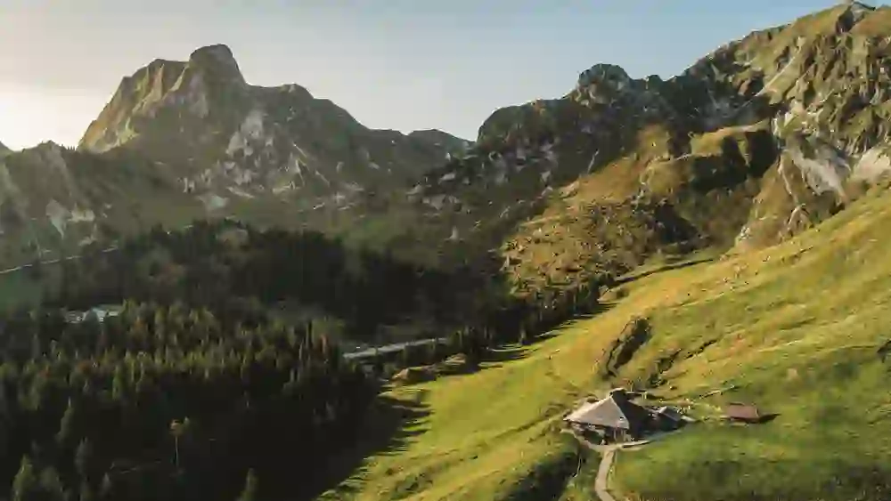 Auf diesem Bild ist erneut die Alp Bire im Naturpark Gantrisch zu sehen – diesmal aus leicht erhöhter Perspektive. Die urige Alphütte liegt in sanftem Licht auf einem grünen Hang, eingerahmt von mächtigen Bergflanken und steilen Felswänden. Im Hintergrund ragt der charakteristische Gipfel des Gantrisch kraftvoll in den Himmel, während dichte Wälder die tiefer gelegenen Täler füllen. Die Szene strahlt stille Erhabenheit, Ursprünglichkeit und die ruhige Kraft der alpinen Natur aus – ein Ort der Einkehr und Weite.