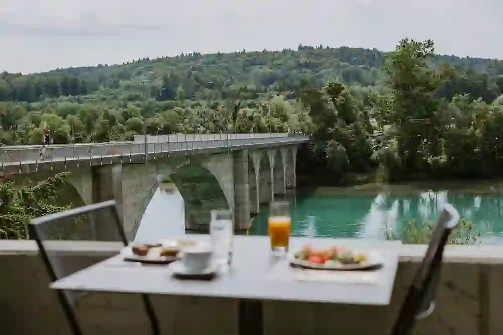 La photo montre une table dressée sur la terrasse du Ristorante-Bar Lago à Hinterkappelen, prête pour un brunch avec vue sur le pont pittoresque et les eaux calmes du lac Wohlensee. Le repas a l'air accueillant et promet un plaisir avec vue. Le décor verdoyant et le reflet dans l'eau créent une ambiance paisible, idéale pour des moments de détente. Ici, les hôtes peuvent profiter de la beauté de la nature tout en se faisant plaisir sur le plan culinaire.