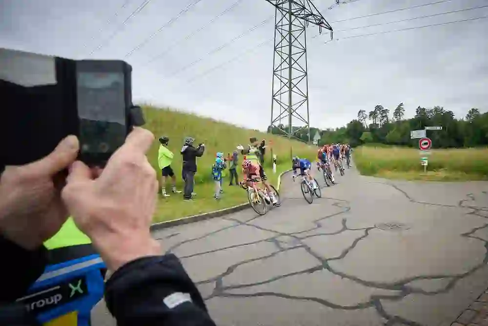 The leading group sweeps through the landscape at breakneck speed, cheered on by cheering fans at the side of the road. The dynamics of the race are palpable - speed, excitement and enthusiasm merge into a perfect sporting moment.