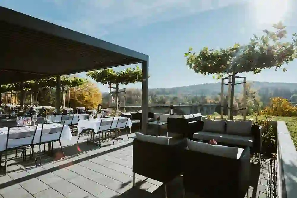 Das Bild zeigt eine geräumige Terrasse mit stilvoll gedeckten Tischen und gemütlichen Loungemöbeln unter einem modernen Pergola-Dach, das einen malerischen Blick auf eine herbstliche Landschaft und eine historische Brücke bietet. Die Atmosphäre wirkt einladend und entspannt, ideal für ein sonniges Herbstessen im Freien.