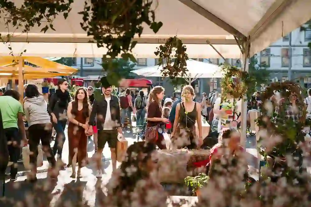 L'immagine mostra un mercato vivace con molte persone. In primo piano ci sono piante e decorazioni, incorniciate da una tenda bianca. Le persone passeggiano tra le bancarelle, alcune portando con sé delle borse. Il tempo è soleggiato e l'atmosfera è allegra e rilassata.