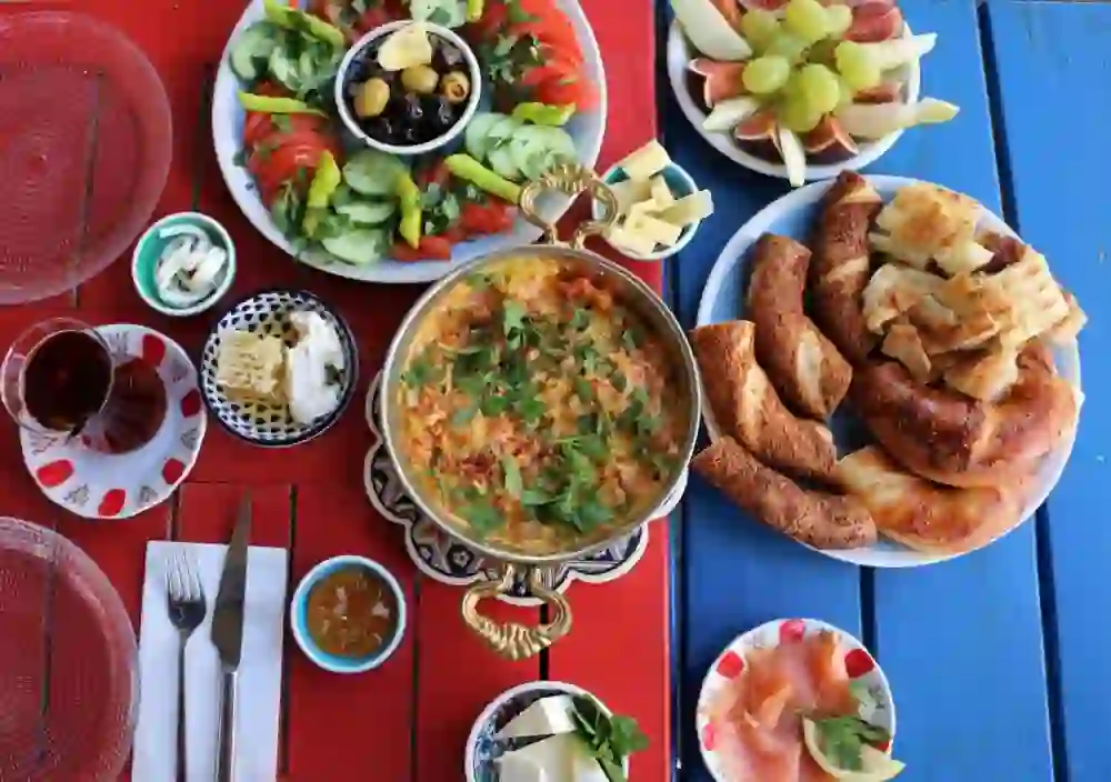 Das Bild zeigt ein farbenfrohes und herzhaftes Frühstück, arrangiert auf einem blau und rot gestreiften Tisch. Es umfasst ein großes, würzig aussehendes Eiergericht in der Mitte, eine Platte mit Brot und Sesamringen, frisches Gemüse, schwarze und grüne Oliven, Käse, Honigwabe, geschnittenen Lachs, sowie Trauben und Feigen. Dazu gibt es Tee und verschiedene Saucen.