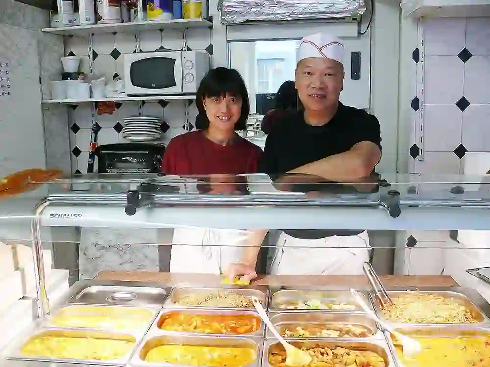 Two friendly chefs smile behind a counter with a variety of hot dishes. The warm and inviting atmosphere of the small restaurant conveys a feeling of home and enjoyment, while the delicious dishes attract guests.