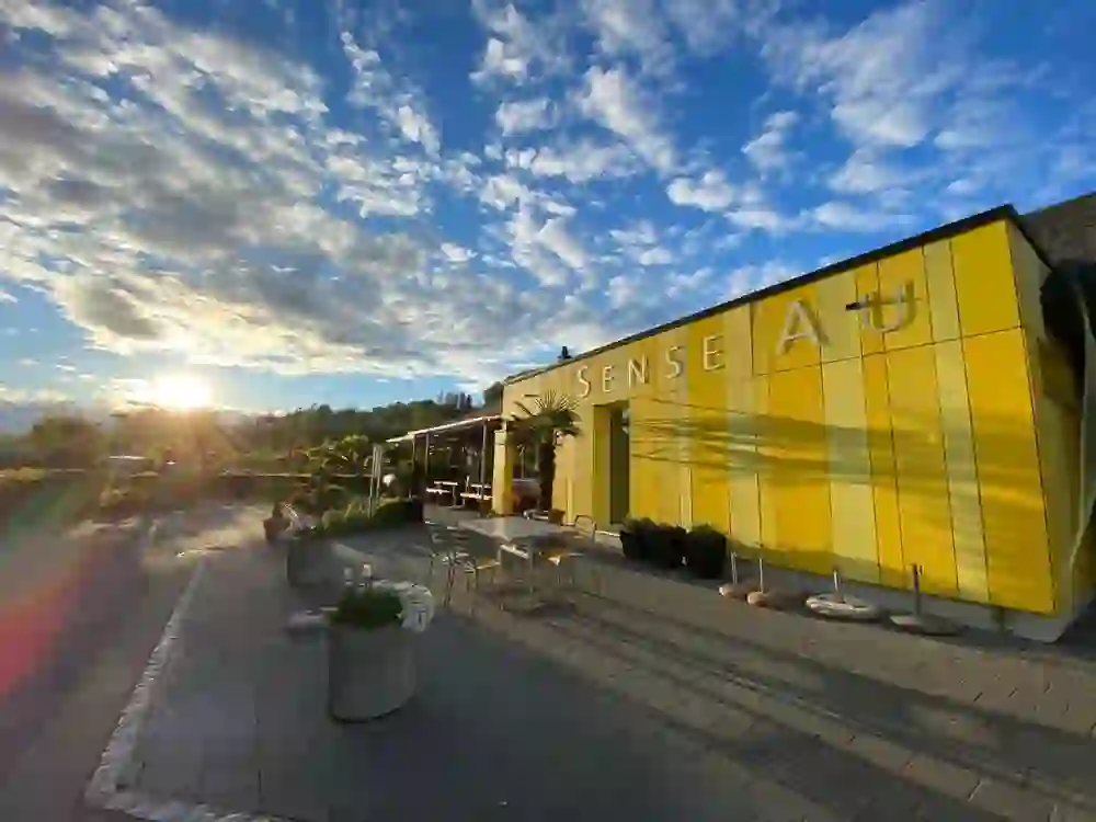 L'image un bâtiment jaune du camping Thörishaus. En arrière-plan, on voit un beau coucher de soleil. La terrasse du jardin est équipée de chaises et de tables et décorée de beaux pots de fleurs.