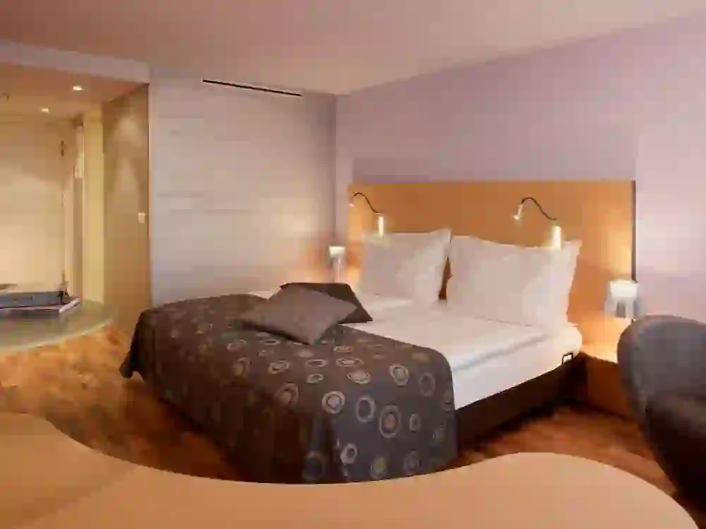 The picture shows a hotel room at the Best Western Plus Hotel, modern and stylishly furnished with a large bed with a circular pattern on the bedspread. The lighting is soft and creates a calming and relaxing atmosphere. Lettering on the wall adds an artistic touch to the room. It appears to be a comfortable place for rest and relaxation.