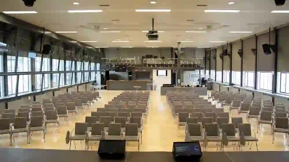 Une grande salle lumineuse avec des rangées de chaises, prête à accueillir un événement. L'atmosphère calme et claire attend la vie et l'activité.