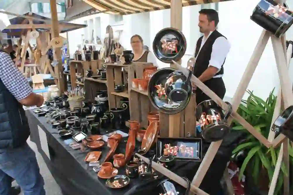 La photo montre un stand sur un marché en plein air où deux vendeurs proposent des produits en céramique faits main. L'ambiance est animée et conviviale, avec un accent sur l'artisanat traditionnel. L'étalage est varié et coloré.