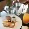 L'image montre un petit-déjeuner agréablement présenté dans un restaurant. Plusieurs crêpes dorées au four, légèrement saupoudrées de sucre glace, sont disposées sur une assiette blanche. À côté se trouve un petit pot de sirop d'érable. Un verre de jus d'orange et un set de couverts plié sur une serviette sont également visibles. En arrière-plan se trouve un vase avec des renoncules roses, et l'on distingue également des salières et des poivrières. La scène est accueillante et appétissante, parfaite pour un petit-déjeuner ou un brunch décontracté.