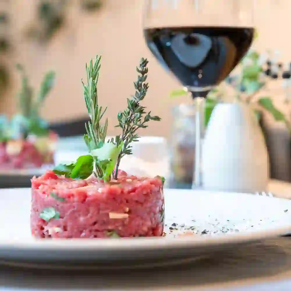 Sur l'image, on voit un tartare artistiquement dressé sur une assiette, décoré d'herbes fraîches. Un verre de vin rouge est posé à côté. La scène dégage une atmosphère élégante et savoureuse, parfaite pour un dîner raffiné dans une ambiance stylée.