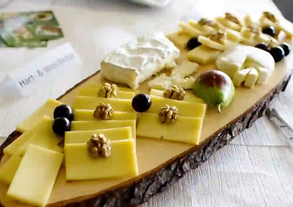 Auf dem Bild ist eine Käseplatte zu sehen, die auf einem rustikalen Holzbrett angerichtet ist. Es gibt verschiedene Sorten von Käse, unter anderem Hartkäse mit Walnüssen und dunkle Trauben als Beilage. Auch ein Stück Weichkäse und eine frische Feige sind zu erkennen. Ein Schildchen mit der Aufschrift „Hart- & Weichkäse“ weist auf die Käseauswahl hin. Die Präsentation ist einfach und ansprechend.
