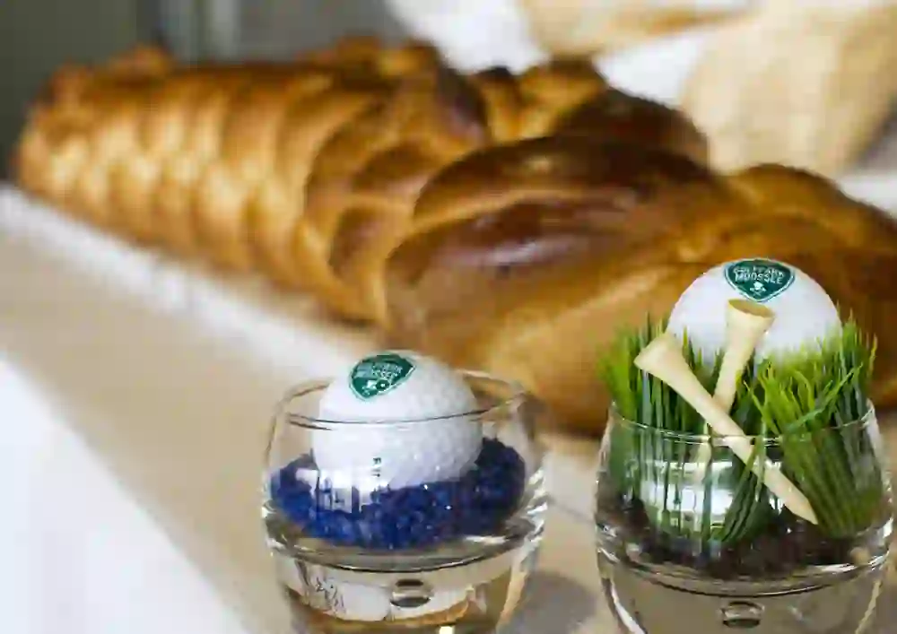 Das Foto zeigt ein großes, geflochtenes Brot im Hintergrund und im Vordergrund zwei Glasvasen mit dekorativen Elementen, die an Golf erinnern. In einer Vase sind ein Golfball auf einem Tee und blaue Dekosteine, in der anderen Vase befinden sich Golftrophäen.