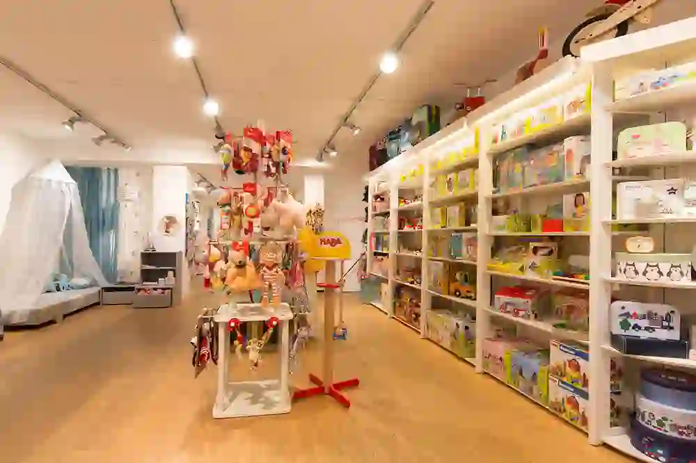 Das Bild zeigt das Innere eines Kinderladens oder Spielzeuggeschäfts, prall gefüllt mit bunten Spielwaren und Kinderartikeln. An den weißen Regalen sind verschiedene Spielzeuge, Bücher und Spiele sichtbar, alles akkurat angeordnet und präsentiert. Im Vordergrund ist ein Stand mit hängenden Stofftieren und Spielsachen. Die Einrichtung ist hell und kinderfreundlich gestaltet, mit einem warmen Holzboden und guter Beleuchtung.