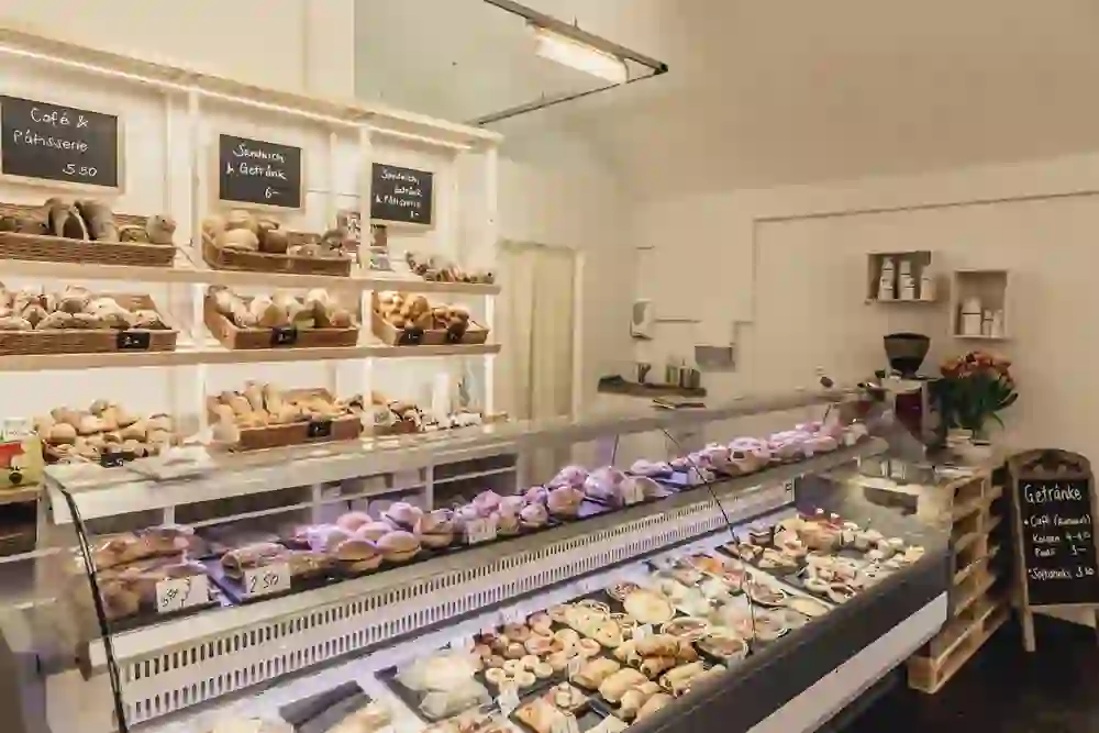 In einer gemütlichen Bäckerei sind Regale mit einer Vielzahl von frischen Broten und Gebäck zu sehen. Die Vitrine ist gefüllt mit verlockenden Patisserien. Die warme Atmosphäre lädt zum Verweilen und Genießen ein, während der Duft von frisch gebackenem Brot die Luft erfüllt.