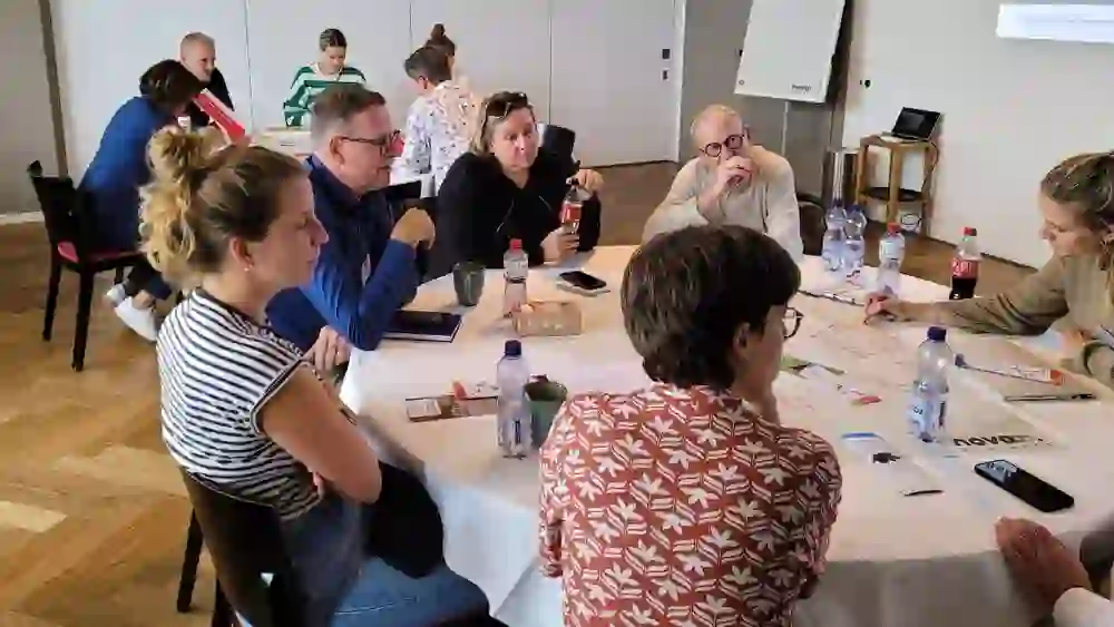 Das Bild zeigt eine Gruppe von Teilnehmern, die intensiv an einem runden Tisch arbeiten. Sie diskutieren und notieren Ideen auf einem großen Blatt Papier. Die Atmosphäre wirkt konzentriert und produktiv, während verschiedene Materialien und Getränke auf dem Tisch verteilt sind. Die lockere Sitzordnung und die entspannte Körperhaltung der Teilnehmer vermitteln eine offene und kollaborative Arbeitsumgebung.
