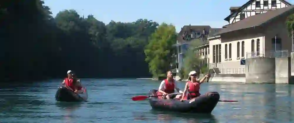 Auf dem Bild paddeln zwei Schlauchboote gemächlich auf der Aare entlang. Die Insassen in leuchtend roten Schwimmwesten geniessen die Fahrt und die Aussicht auf das Ufer mit seiner dichten Bewaldung und einem historischen Gebäude. Es ist ein friedlicher Tag auf dem Wasser, voller Entspannung und gemeinschaftlicher Freude.