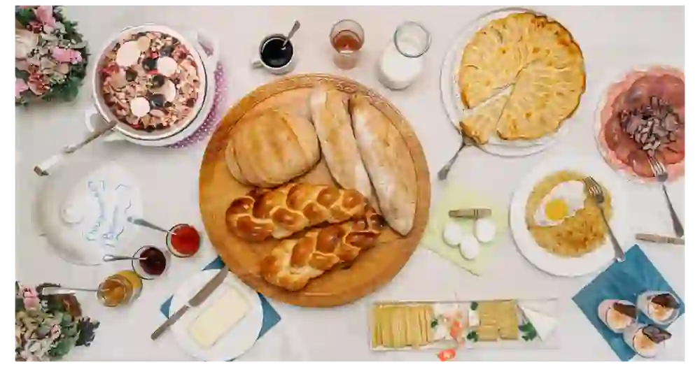 Auf dem Bild ist ein üppig gedeckter Frühstückstisch zu sehen. Zu den Speisen gehören verschiedene Brotarten, darunter ein Zopfbrot, sowie eine Schüssel Müsli mit frischen Beeren. Weiterhin gibt es eine Tarte, Aufschnitt, darunter Schinken und Salami, und Rührei mit einem Spiegelei. Dazu gibt es Beilagen wie Butter, Honig, Marmelade und Käse, sowie Getränke wie Milch und Kaffee.