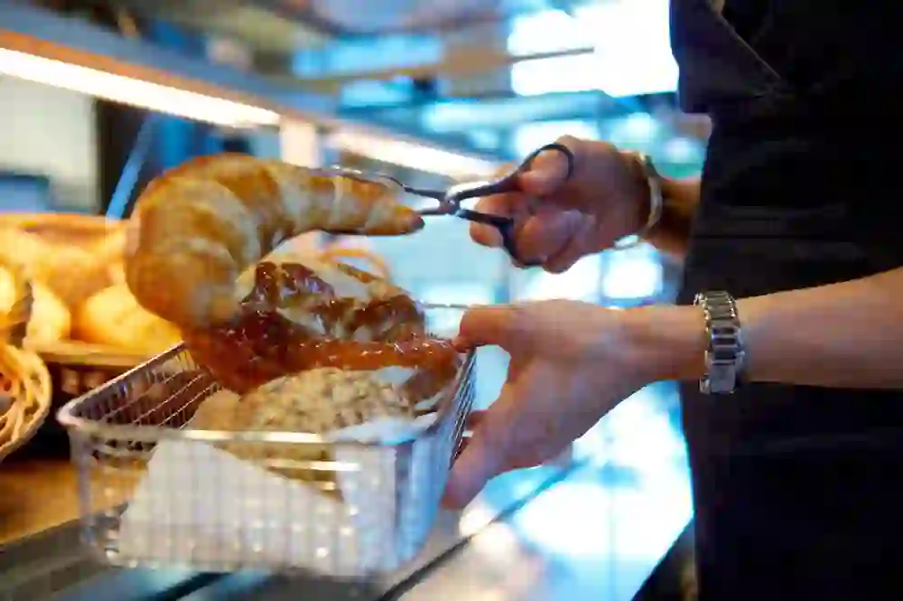 Das Bild zeigt eine Person, die mit einer Zange ein frisch gebackenes Croissant aus einem Korb nimmt. Im Hintergrund sind weitere Backwaren zu sehen, was auf eine Bäckerei oder ein Café hinweist. Die Person trägt eine Armbanduhr, was auf eine Detailaufnahme im Rahmen eines Frühstücks oder einer Kaffeepause deutet.