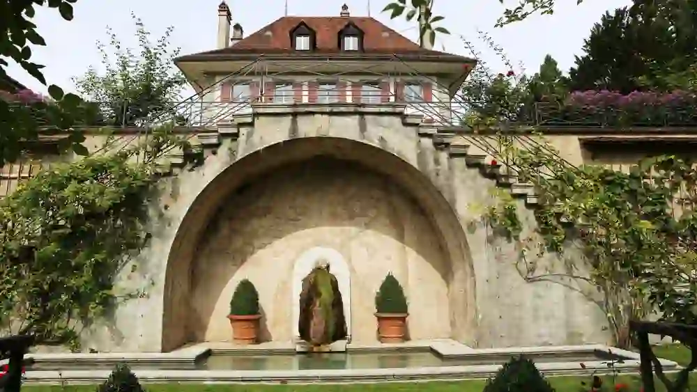 Auf dem Bild ist ein historischer Garten mit einem großen, gewölbten Wasserspiel zu sehen. Über dem Wasserspiel befindet sich ein Gebäude mit einem Balkon und rot-weißen Fensterläden. Der Garten ist gepflegt, mit sorgfältig beschnittenen Hecken und einem sauber geschnittenen Rasen. Bunte Blumen und kletternde Pflanzen zieren das Mauerwerk, und das Arrangement bietet einen ruhigen Ort der Entspannung.