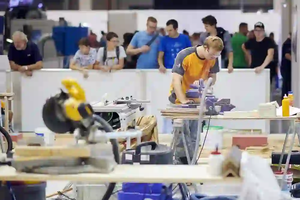 Das Bild zeigt eine Person, die an einem Werkstück arbeitet, umgeben von Werkzeugen und Maschinen, während Zuschauer bei einer Veranstaltung wie den SwissSkills im Hintergrund zusehen.