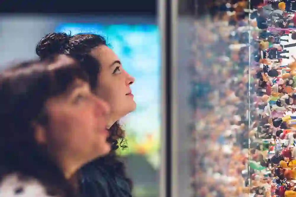 Zwei Besucherinnen betrachten fasziniert eine dichte Wand aus unzähligen Figuren in der Ausstellung «Massen». Ihre neugierigen Blicke spiegeln Staunen und spielerische Entdeckerlust – ein Moment, in dem Vielfalt und Überfluss lebendig werden.