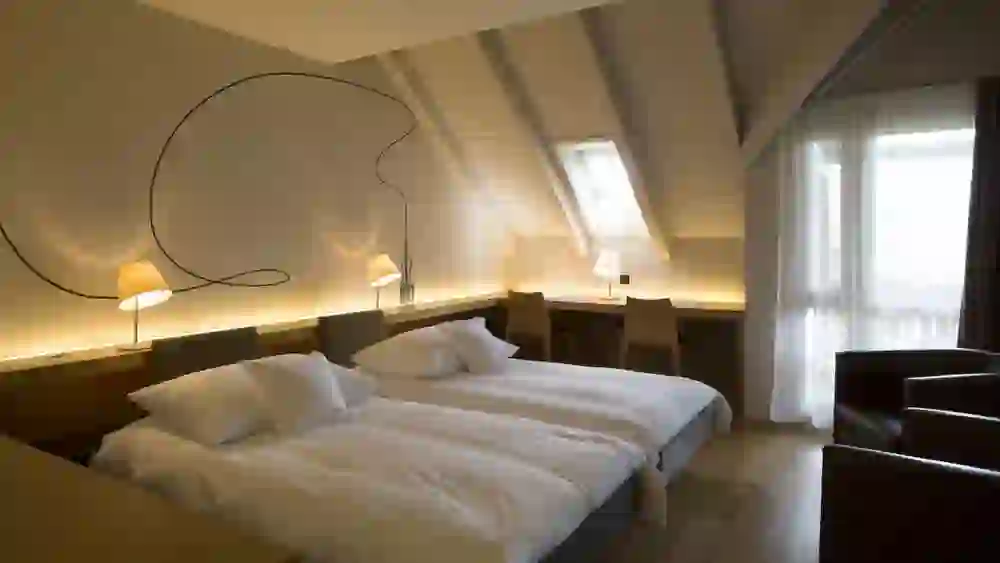 Das Bild zeigt ein modernes Hotelzimmer mit zwei Einzelbetten. Über den Betten befindet sich eine interessante Wandbeleuchtung, die eine große, abstrakte Form an die Wand wirft. Es gibt zwei Tischlampen und eine lange, hinterleuchtete Ablage, die den Raum in ein warmes Licht taucht. Die Dachschrägen und das Dachfenster verleihen dem Raum eine gemütliche Atmosphäre, und es gibt Sitzgelegenheiten sowie einen Schreibtisch am Fenster.