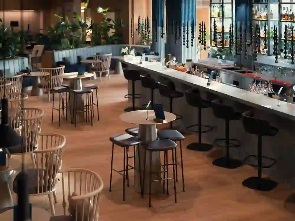 Un espace bar et lounge stylé et moderne, aux tons chauds de bois et à la décoration élégante. L'atmosphère est détendue et sophistiquée, idéale pour des rencontres conviviales ou des discussions tranquilles. Un sentiment de confort, de modernité et d'ambiance urbaine imprègne l'espace.
