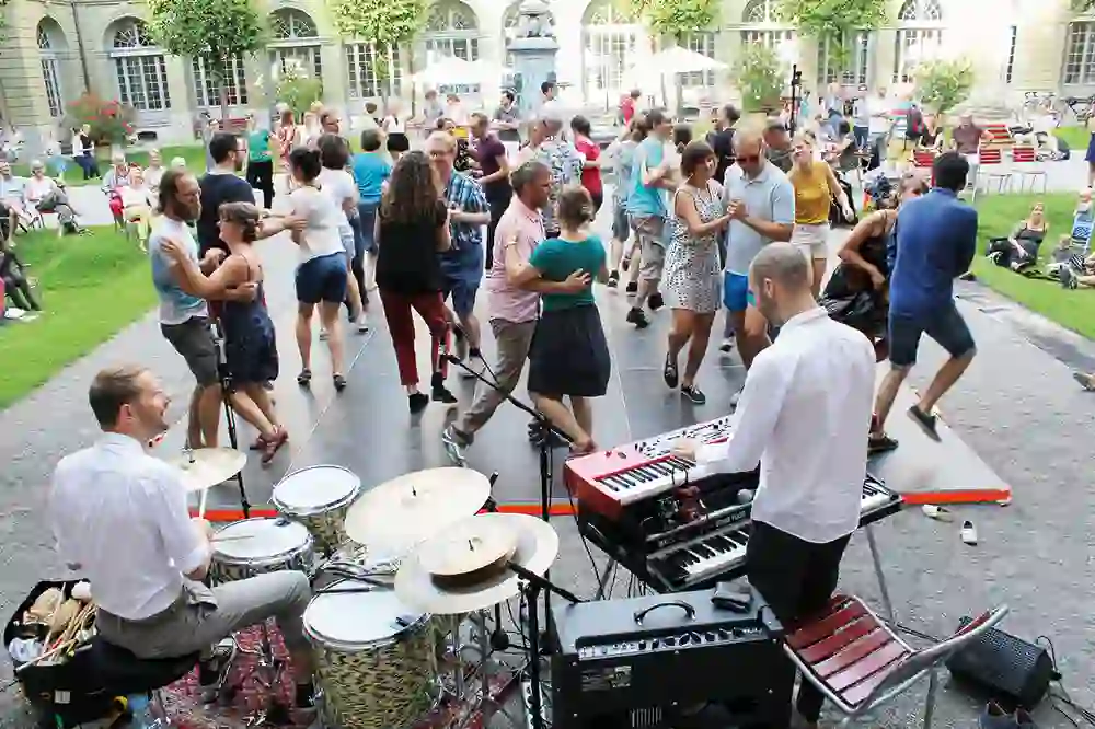 Auf dem Bild ist eine fröhliche Menschenmenge beim Tanzen in einem Open-Air-Bereich des Berner Generationenhauses zu sehen. Musiker untermalen das Geschehen, während die Stimmung ausgelassen und heiter ist, eine Gemeinschaft, die gemeinsam den Rhythmus und das Beisammensein geniesst.