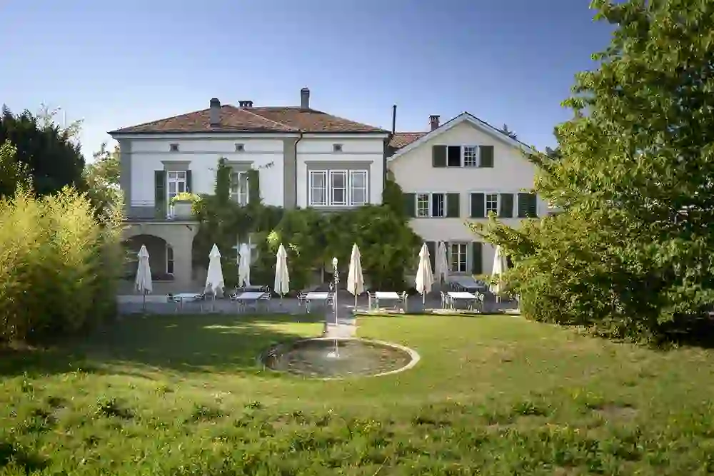 Auf dem Bild ist das Restaurant Schöngrün zu sehen, dessen Aussenterrasse mit weissen Sonnenschirmen eine idyllische und friedliche Atmosphäre ausstrahlt. Die umgebende grüne Landschaft und der Brunnen vermitteln eine Oase der Ruhe.