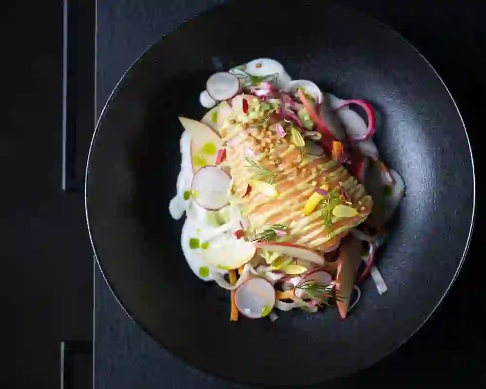 Piatto colorato con tenero filetto di salmone su insalata di verdure, guarnito con semi di senape, fiori ed erbe in una ciotola nera.