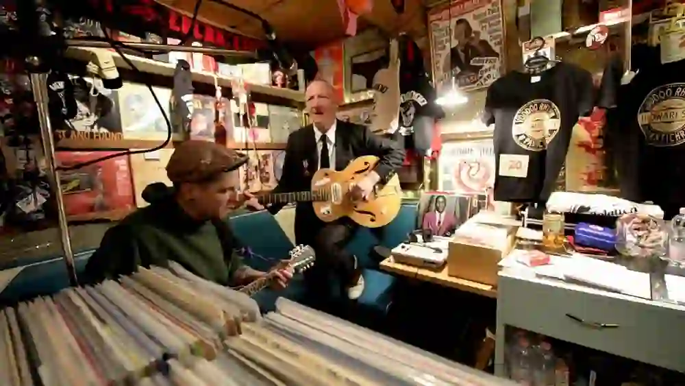 Das Bild zeigt zwei Personen in einem lebhaft dekorierten Raum, vermutlich in einem Musikladen oder einem ähnlichen Geschäft, das mit Musik und Kultur verbunden ist. Eine Person spielt Gitarre, während die andere, in Anzug und Krawatte gekleidet, ebenfalls eine Gitarre hält. Der Raum ist voller Musik- und Kulturartikel, darunter Schallplatten, Poster, T-Shirts und andere Fanartikel. Die Atmosphäre wirkt entspannt und von einer Leidenschaft für Musik geprägt.