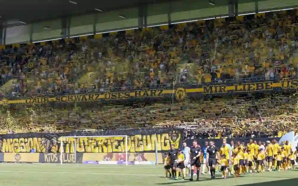 Auf dem Bild ist eine Szene vor Beginn eines Fußballspiels im Stadion des BSC Young Boys zu sehen. Im Vordergrund sind die Spieler und das Schiedsrichterteam zu erkennen, die sich zur Begrüßung oder für ein Mannschaftsfoto aufstellen. Im Hintergrund ist eine lebendige Zuschauermenge in einer Kurve des Stadions zu sehen, viele davon in den Vereinsfarben Gelb und Schwarz