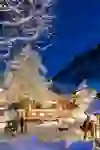 Snow-covered village at night with illuminated wooden houses, trees and a mountain backdrop. Warm lights create a cosy, festive atmosphere.