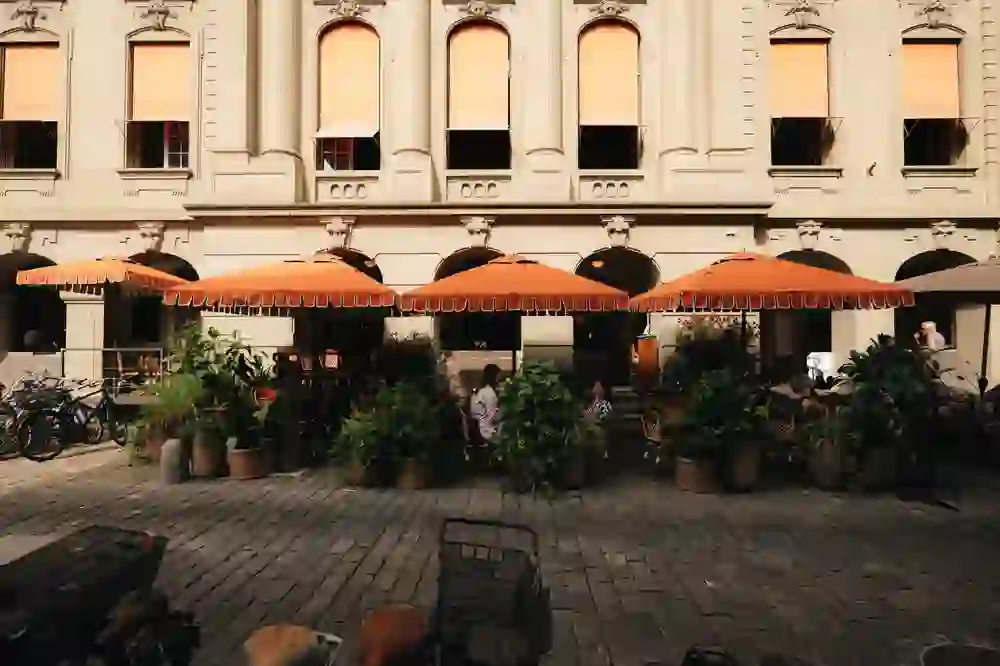 Accogliente terrazza con ombrelloni arancioni di fronte a una facciata storica. Piante rigogliose e sedute informali creano un ambiente invitante per conversazioni rilassate nel cuore della città.