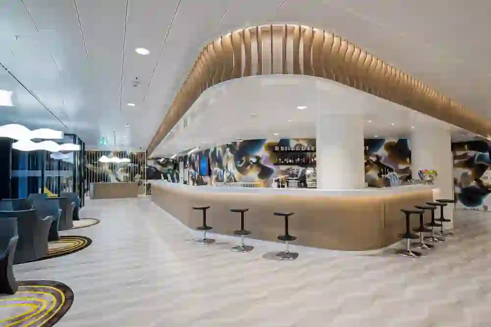 Das Bild zeigt das Innere einer modernen Hotellobby oder Lounge mit einer geschwungenen Bar im Zentrum. Die Einrichtung ist futuristisch, mit wellenförmigen Mustern auf dem Boden und einer dynamischen, abstrakten Wandgestaltung. Die Möbel sind stilvoll und zeitgemäß, und die Beleuchtung ist kunstvoll und sorgt für eine lebendige, einladende Atmosphäre. Es ist ein Raum, der Modernität und Design betont und den Besuchern ein Gefühl von Eleganz und Trendbewusstsein vermittelt.