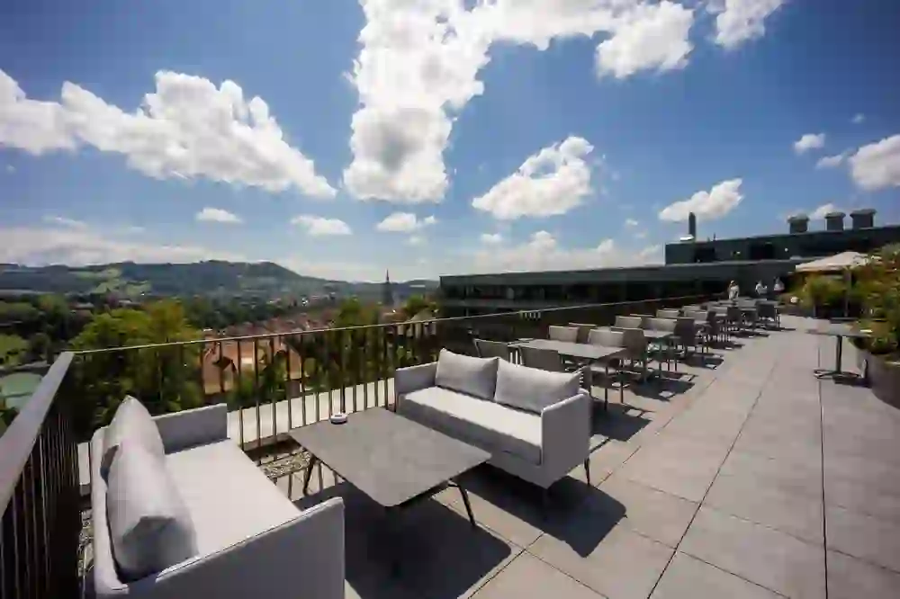 Das Bild zeigt eine Dachterrasse mit moderner Lounge-Möblierung und einer atemberaubenden Aussicht auf eine Hügellandschaft und die Stadtarchitektur im Hintergrund. Der Himmel ist klar mit einigen Schönwetterwolken, was eine offene und beruhigende Atmosphäre schafft. Die Terrasse ist ein idealer Ort für Entspannung und soziale Zusammenkünfte, mit einem Gefühl der Freiheit und Erhabenheit über der städtischen Umgebung von der Stadt Bern.