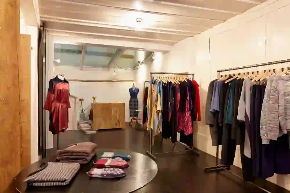 Das Bild zeigt das Innere eines modischen Bekleidungsgeschäfts. An mehreren Kleiderständern sind sorgfältig ausgewählte Kleidungsstücke präsentiert. Auf einem runden Tisch in der Mitte sind gefaltete Textilien platziert. Die Beleuchtung ist warm, und durch ein Oberlicht kommt Tageslicht herein. Der Raum hat einen modernen, minimalistischen Stil mit klaren Linien und einer Kombination aus natürlichen Materialien wie Holz und Beton.