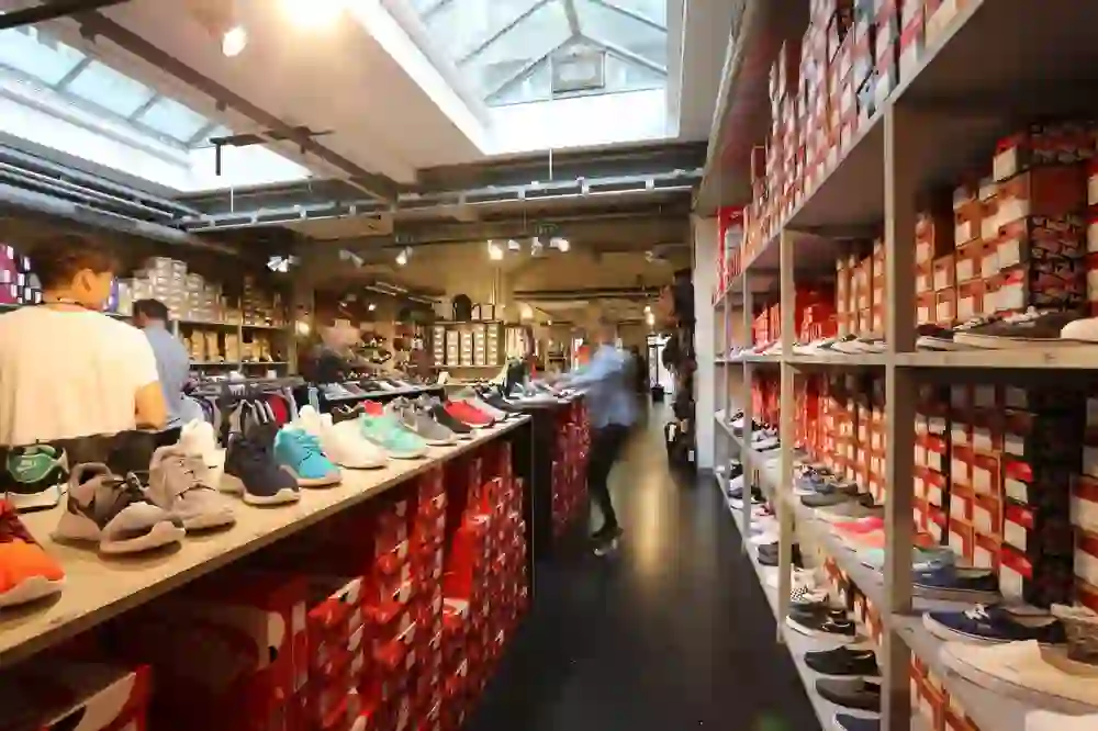 Das Bild zeigt das Innere eines Schuhgeschäfts mit einer breiten Auswahl an Sportschuhen verschiedener Marken, die auf Regalen präsentiert werden. Die Schuhe sind ordentlich neben den passenden Schachteln platziert. Das Geschäft hat eine industrielle Atmosphäre mit sichtbaren Rohren an der Decke und einem Oberlicht, das für eine helle Beleuchtung sorgt. Im Hintergrund sind weitere Modeartikel zu erkennen. Kunden und Mitarbeiter sind teilweise sichtbar.